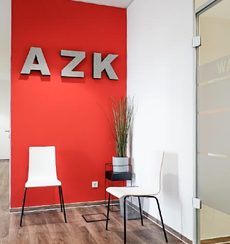 Zwei weiße Stühle stehen vor einer roten Wand mit dem Schriftzug "AZK" und einer Pflanze auf einem Tisch.