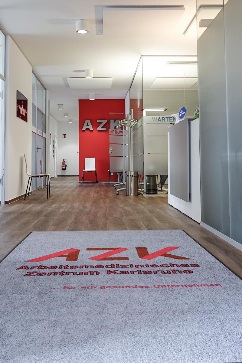 Ein moderner Flur mit einem Teppich, auf dem "AZK Arbeitsmedizinisches Zentrum Karlsruhe" steht, führt zu einer Wartebereichstür.