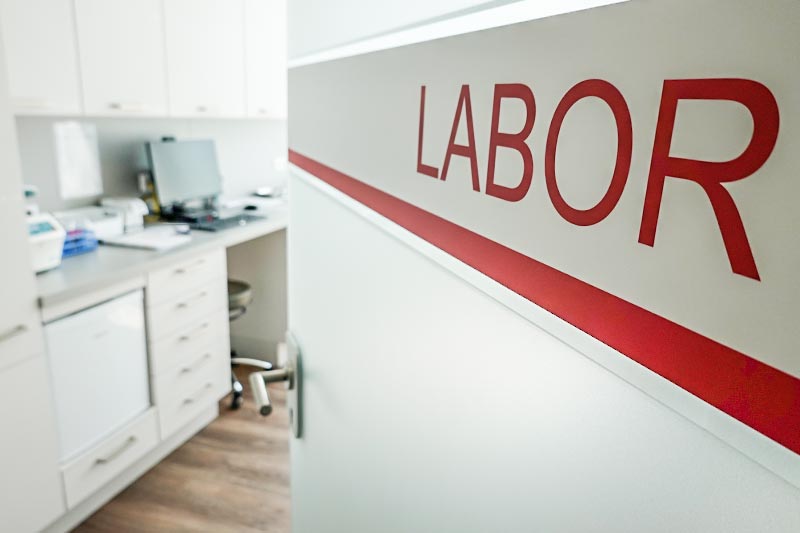 Ein Laborraum mit einem weißen Schrank, auf dem in roter Schrift "LABOR" steht, und einem Schreibtisch im Hintergrund.